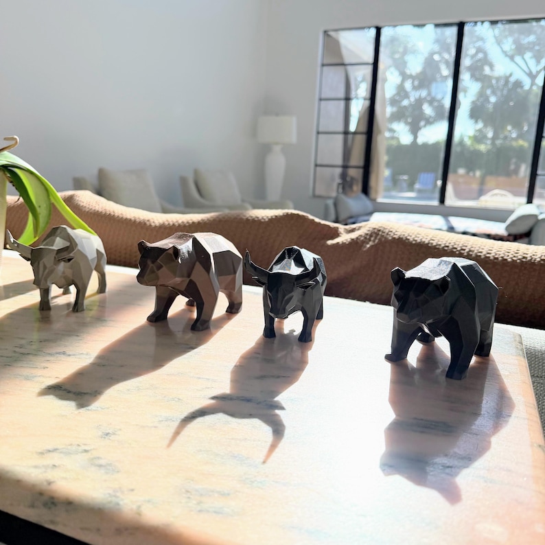 Four geometric animal statues, two bulls and two bears, in light gray, bronze, black, and dark gray, are lined up on a marble surface in a sunlit room.
