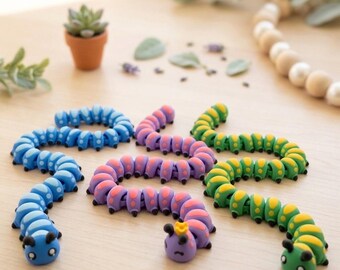 Puede incluir: Una colección de coloridas gomas de borrar con forma de oruga en azul, morado y verde. Cada goma presenta un cuerpo segmentado y una cara linda con pequeñas antenas. Una pequeña planta suculenta en maceta está en el fondo.