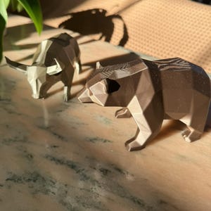 A bronze geometric bear statue is in the foreground, with a light gray geometric bull statue slightly out of focus behind it, on a marble surface.