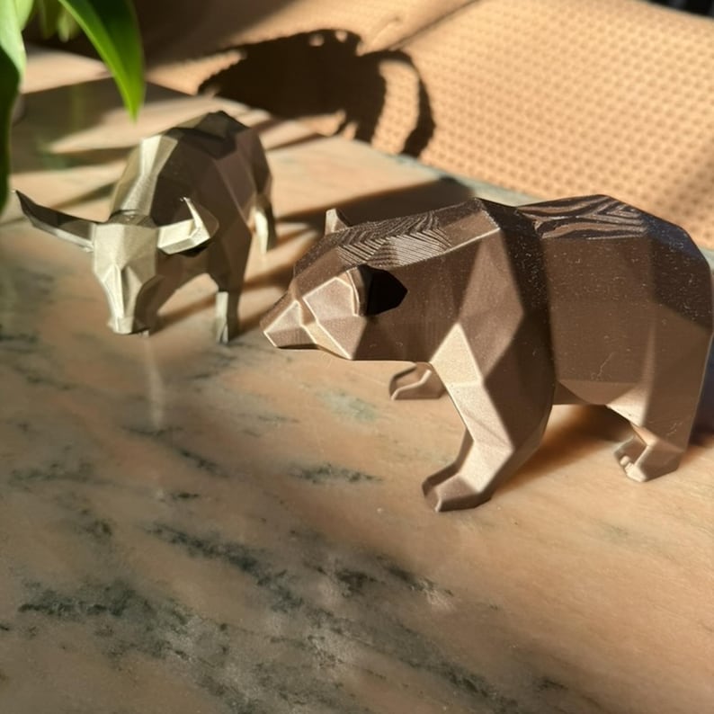 A bronze geometric bear statue is in the foreground, with a light gray geometric bull statue slightly out of focus behind it, on a marble surface.