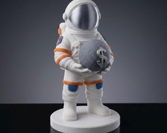 Puede incluir: Figura de astronauta blanca con detalles en naranja y azul, sosteniendo una esfera gris con un signo de dólar plateado. El astronauta tiene un casco plateado reflectante y está sobre una base circular blanca. Artículo decorativo.