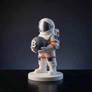 May include: A white astronaut figurine holding a dark grey sphere with a Bitcoin symbol. The astronaut suit has orange and blue accents. The figurine stands on a white circular base against a dark background.