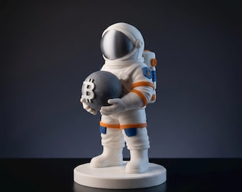 Puede incluir: Una figura de astronauta blanca sosteniendo una esfera gris oscuro con un símbolo de Bitcoin. El traje de astronauta tiene detalles en naranja y azul. La figura se encuentra sobre una base circular blanca sobre un fondo oscuro.