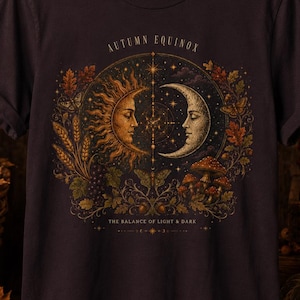May include: A dark brown short-sleeve t-shirt featuring a graphic print of a sun and moon face surrounded by autumn leaves, wheat, and mushrooms. The text reads AUTUMN EQUINOX and THE BALANCE OF LIGHT & DARK in a gold-toned, ornate font style.