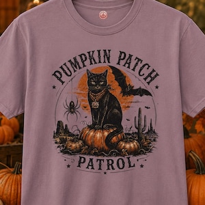May include: A mauve short-sleeve t-shirt featuring a graphic print of a black cat sitting on a pumpkin. The text PUMPKIN PATCH PATROL surrounds the cat, which is accented by a spider, a bat, and a desert landscape with a large orange moon.