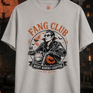 May include: A light gray short-sleeve t-shirt featuring a graphic of a vampire on a motorcycle. The text reads FANG CLUB, NIGHT RIDERS CHAPTER, and EST. 1897. The design includes a full moon, bats, and orange accents on a circular background.