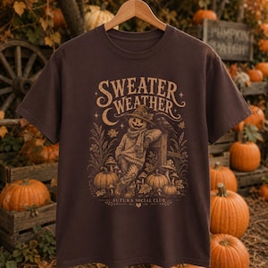 May include: A dark brown short-sleeve t-shirt featuring a tan graphic print of a pumpkin-headed figure wearing a sweater and hat, surrounded by pumpkins, mushrooms, and a raven. The text reads "SWEATER WEATHER" and "AUTUMN SOCIAL CLUB."