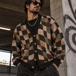 May include: A long-sleeve button-up cardigan featuring a distorted checkerboard pattern in shades of brown, tan, olive green, and black. The garment has a solid black V-neck placket, black ribbed cuffs, a black hem, and five circular buttons.