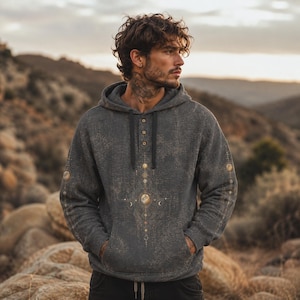 May include: A charcoal gray long-sleeve hoodie featuring a textured fabric finish, a three-button placket with a drawstring hood, and a central vertical graphic design composed of celestial symbols, moons, and stars in muted gold and white tones.