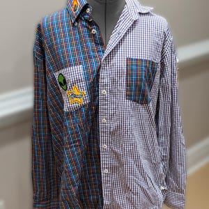 May include: A button-down shirt with a split design. One side features a plaid pattern in blue, red, and yellow, while the other has a white and black gingham check. Patches with an alien and the word "Space" are sewn on the plaid side.