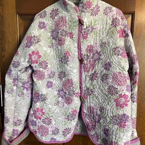 May include: A quilted jacket with a floral pattern in shades of pink, purple, and green on a light gray background. The jacket has a mandarin collar, wooden toggle closures, and a contrasting pink trim.