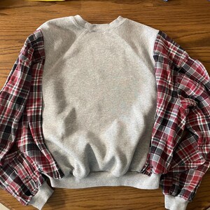 May include: A gray sweatshirt with long sleeves featuring a red, black, and white plaid pattern. The sleeves are a contrasting fabric, adding a unique design element to the casual top. The cuffs and neckline are ribbed.