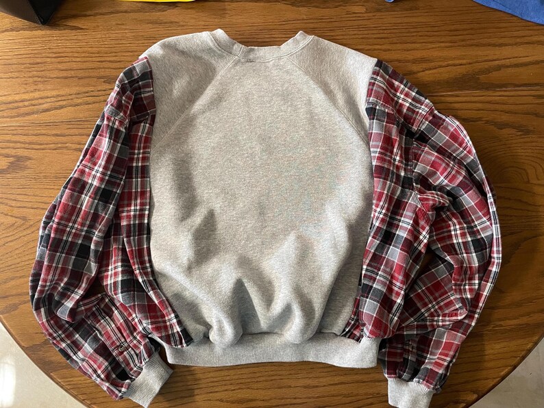 May include: A gray sweatshirt with long sleeves featuring a red, black, and white plaid pattern. The sleeves are a contrasting fabric, adding a unique design element to the casual top. The cuffs and neckline are ribbed.