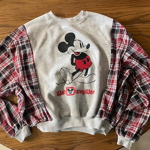 May include: Gray sweatshirt with Mickey Mouse graphic and red, black, and white plaid sleeves. The graphic features Mickey Mouse in a red and black outfit. The text "Walt Disney Wor" is below the graphic. The sweatshirt has a crew neck and ribbed cuffs.