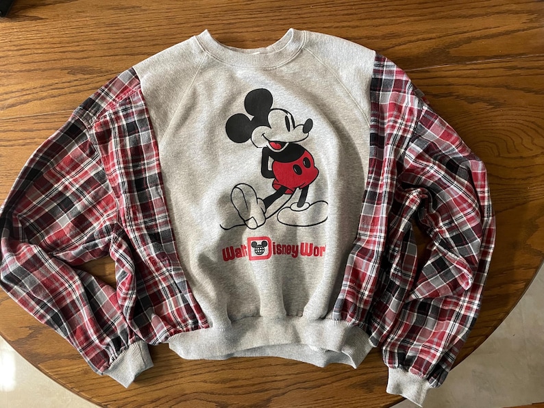 May include: Gray sweatshirt with Mickey Mouse graphic and red, black, and white plaid sleeves. The graphic features Mickey Mouse in a red and black outfit. The text "Walt Disney Wor" is below the graphic. The sweatshirt has a crew neck and ribbed cuffs.