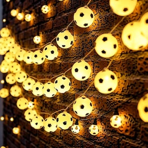 May include: String lights featuring soccer ball-shaped bulbs. The bulbs are white with black pentagon shapes, emitting a warm yellow glow. The lights are strung against a textured brick wall, creating a festive atmosphere.