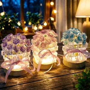 May include: Three decorative floral lamps with a warm glow. Each lamp features a cluster of roses in purple, pink, and blue atop a clear, cube-shaped base. A pink ribbon is tied around the base. The lamps sit on a wooden surface.