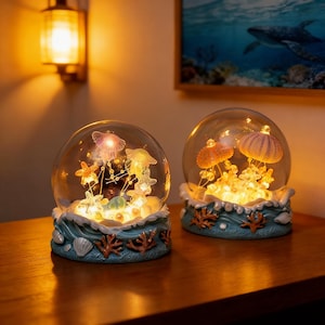 May include: Two decorative glass globes with illuminated jellyfish and starfish inside. The globes sit on a blue base with coral and seashell accents. The globes are on a wooden surface, with a wall sconce and a framed picture in the background.