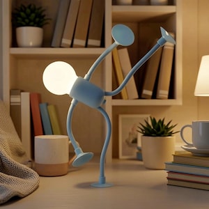 Może przedstawiać: Jasnoniebieska lampa biurkowa o unikalnym, stylizowanym designie, przypominającym postać w ruchu. Lampa posiada dużą, okrągłą żarówkę i elastyczne ramiona zakończone okrągłymi i płaskimi dyskami. Na biurku z książkami i małą rośliną.