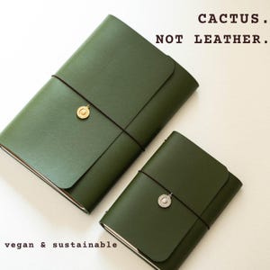 Może przedstawiać: Dwa oliwkowozielone notatniki z gładkimi okładkami, zabezpieczone cienkimi czarnymi gumkami i okrągłymi metalowymi zawieszkami. Tekst CACTUS. NOT LEATHER. oraz vegan & sustainable jest wydrukowany na czarno. Notatniki mają zapięcie na klapkę.