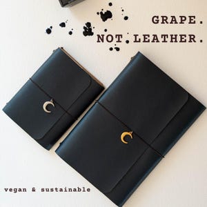 Peut inclure: Deux carnets noirs à couverture lisse, chacun maintenu par un fin élastique sombre orné d'un pendentif en forme de croissant de lune. Le petit carnet a une breloque argentée, le grand une dorée. Texte : GRAPE. NOT LEATHER. vegan & sustainable.