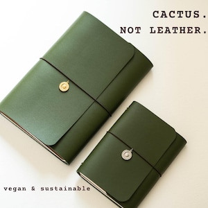May include: Two olive green journals with smooth covers, secured by thin black elastic bands and round metallic charms. The text CACTUS. NOT LEATHER. and vegan & sustainable is printed in black. The journals have a flap closure design.