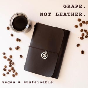 May include: A dark brown journal with a smooth finish, secured by a thin elastic band featuring a silver spiral charm. The text GRAPE. NOT LEATHER. and vegan & sustainable is visible. A glass of black coffee and scattered coffee beans are present.