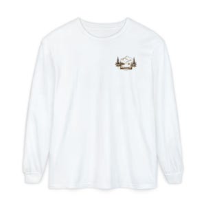 Puede incluir: Una camiseta blanca de manga larga con un pequeño gráfico marrón en la parte superior izquierda del pecho. La ilustración muestra una canoa flotando en el agua frente a una cordillera rodeada de pinos, en un estilo de dibujo lineal simple.