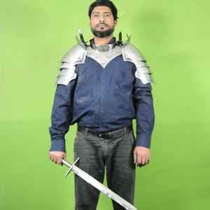 Könnte beinhalten: Eine Person trägt eine silberne Schulterpanzerung aus Metall mit einem blauen Button-Down-Hemd und einer grauen Hose und hält ein Schwert. Die Rüstung hat mehrere geschichtete Platten und dekorative Spitzen. Das Schwert hat eine silberne Klinge und einen silbernen Griff.