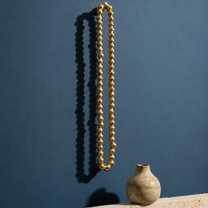 May include: A long, gold-colored beaded chain with a reflective, metallic finish hangs vertically. Below the chain sits a small, round, beige ceramic vase with a narrow neck, positioned on a textured, light-colored stone block surface.