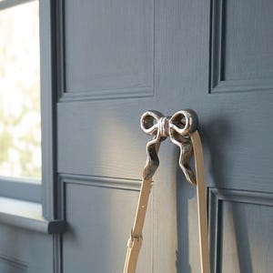 May include: A silver-toned metal wall hook shaped like a bow, mounted on a dark blue paneled wall. A cream-colored handbag with a circular handle and an adjustable shoulder strap hangs from the hook. A small speckled tray with keys sits nearby.