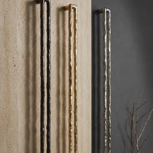 Könnte beinhalten: Drei lange, vertikale Türgriffe mit einer texturierten, organischen und unebenen Oberfläche. Die Griffe sind in einer Reihe angeordnet und weisen ein mattes schwarzes Finish, ein poliertes goldenes Finish und ein metallisch silbernes Finish auf, jeweils mit einem zentralen offenen rechteckigen Schlitz.