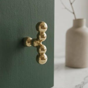 May include: A polished brass cabinet knob featuring a vertical arrangement of four spherical balls attached to a central mounting base, mounted on a dark forest green textured surface. The metal finish is reflective and smooth in texture.