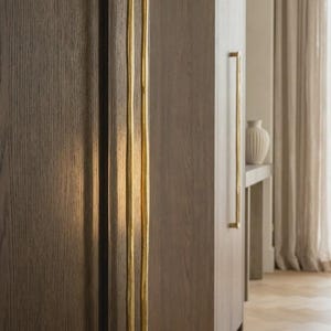 May include: A long, vertical, gold-toned metal door handle with a continuous, rounded, U-shaped design. The handle is mounted onto a dark brown, wood-grained cabinet door. The surface of the metal handle has a slightly textured, organic finish.