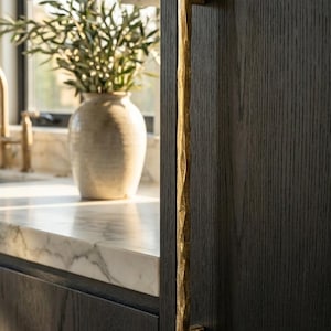 Puede incluir: Un tirador de armario largo y vertical con un acabado dorado martillado y texturizado, fijado a una puerta de armario de madera de color gris carbón oscuro. El tirador presenta un diseño recto y fino con una textura rugosa e irregular.