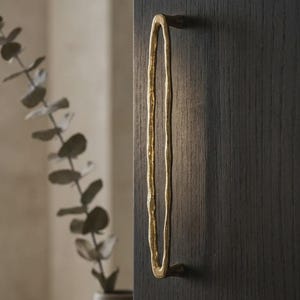 以下が含まれることがあります： 暗い木製のキャビネットドアに取り付けられた、テクスチャ加工された有機的なデザインの金色の細長いキャビネットプル。 プルは垂直方向に配置され、わずかに不規則な形状をしています。 背景には緑のある花瓶があります。