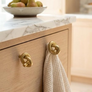 May include: Two circular brass cabinet knobs with a knotted design attached to a light wood drawer front. A cream-colored waffle-textured towel hangs from the right knob, while a bowl of pears sits on the white marble countertop above.