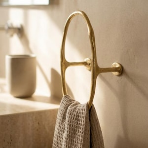 May include: A gold-toned metal towel ring with an irregular, organic oval shape mounted to a wall. The fixture features a textured, matte finish and holds a folded, beige-coloured waffle-weave fabric towel draped through the centre loop.