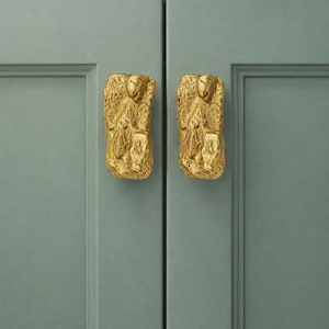 May include: Two rectangular cabinet pulls with a textured, uneven gold finish. Each pull features a raised, abstract relief pattern on the front surface. The pulls are mounted vertically on two adjacent sage green cabinet doors with recessed panels.