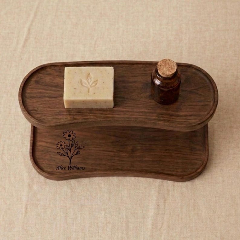 May include: A two-tiered, dark wood tray with an oval top tier and a rectangular bottom tier. The top tier holds a bar of soap with a leaf design and a small brown bottle with a cork stopper. The bottom tier has a floral design and the name "Alice Williams".