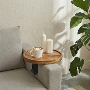 May include: A wooden sofa arm tray with a black metal bracket, holding a white mug with a heart-shaped design in the foam and two white candles. The tray is placed on the arm of a gray sofa.
