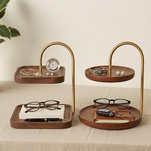 Puede incluir: Dos organizadores de dos niveles con bandejas de madera marr&oacute;n oscuro y marcos de metal dorado curvos. Uno tiene bandejas cuadradas con un reloj peque&ntilde;o, joyas, gafas y un bol&iacute;grafo. El otro tiene bandejas redondas con pulsera, pendientes y llaves.