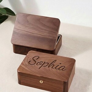 May include: Two wooden boxes with a rich brown finish. One box has its lid open, revealing a lighter interior. The other box is engraved with the name "Sophia" on the lid and features a small gold-colored lock.