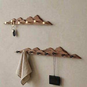 May include: Two wall-mounted wooden coat racks shaped like mountain ranges. The top rack features gold-toned pegs and holds a set of keys. The bottom rack has black pegs and holds a beige fabric cloth and a black card holder on a chain.