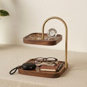 Puede incluir: Una bandeja organizadora de madera de dos niveles con un brazo de soporte curvo de metal dorado. El nivel superior sostiene una pulsera dorada y un peque&ntilde;o reloj redondo. El nivel inferior contiene unas gafas, un estuche de auriculares negro, una libreta y un bol&iacute;grafo.