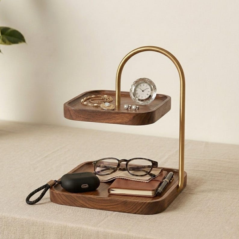 Puede incluir: Una bandeja organizadora de madera de dos niveles con un brazo de soporte curvo de metal dorado. El nivel superior sostiene una pulsera dorada y un peque&ntilde;o reloj redondo. El nivel inferior contiene unas gafas, un estuche de auriculares negro, una libreta y un bol&iacute;grafo.