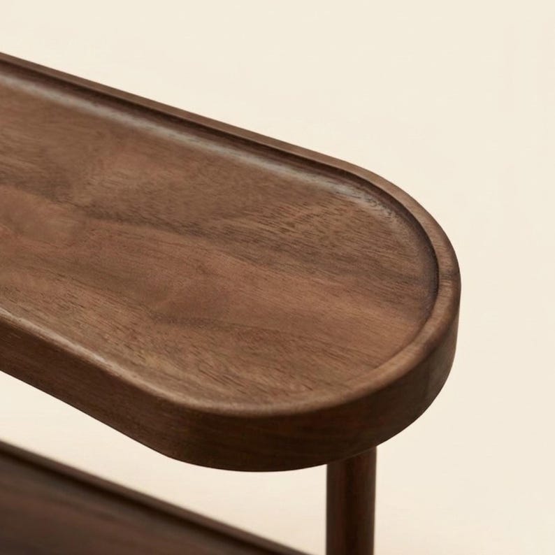 May include: Close-up of a wooden tray with a curved edge and a dark brown finish. The tray is supported by a single, round leg. The wood grain is visible, adding texture to the design. The tray is likely for serving or display.