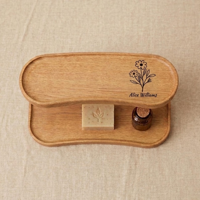 May include: A two-tiered wooden tray with a natural finish. The top tier features a floral design and the name "Alice Williams". A bar of soap with a leaf design and a small glass bottle are on the lower tier.