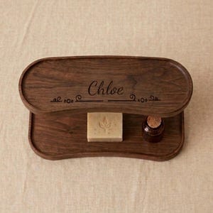 May include: A two-tiered wooden tray with the name "Chloe" engraved on the top tier. The tray holds a bar of soap with a leaf design and a small brown bottle with a cork stopper. The wood has a rich, dark brown color.