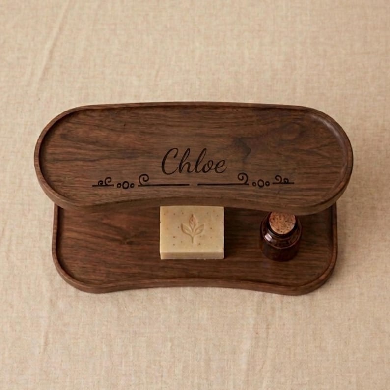 May include: A two-tiered wooden tray with the name "Chloe" engraved on the top tier. The tray holds a bar of soap with a leaf design and a small brown bottle with a cork stopper. The wood has a rich, dark brown color.